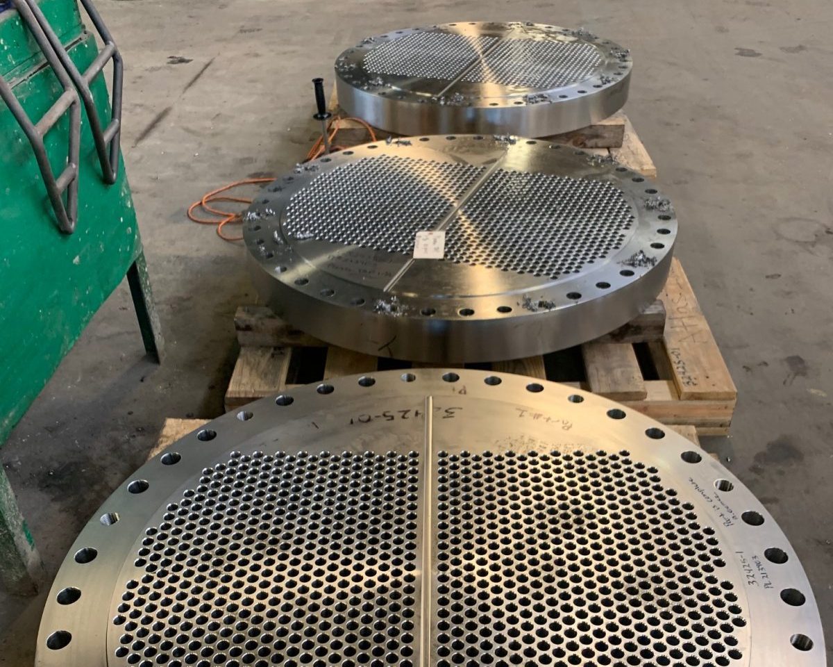 Three large precision-machined tubesheets made from 5-1/4” thick 316LEZ stainless steel, featuring a series of uniformly drilled holes and flanges, placed on wooden pallets in an industrial setting.