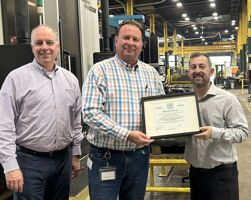 Three men in a factory, with one holding a framed award, standing in front of large industrial machines and yellow beams.