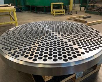 Two images of a large, precision-machined tubesheet made from 5” thick 2205 Duplex Stainless Steel. The tubesheet features a machined beveled edge, raised face, and 692 drilled holes with a 1.514” diameter and two expansion grooves in each hole. The part was plasma cut and machined to a 56.500” OD with precise edge beveling, demonstrating Sandmeyer Steel’s advanced machining and drilling capabilities.